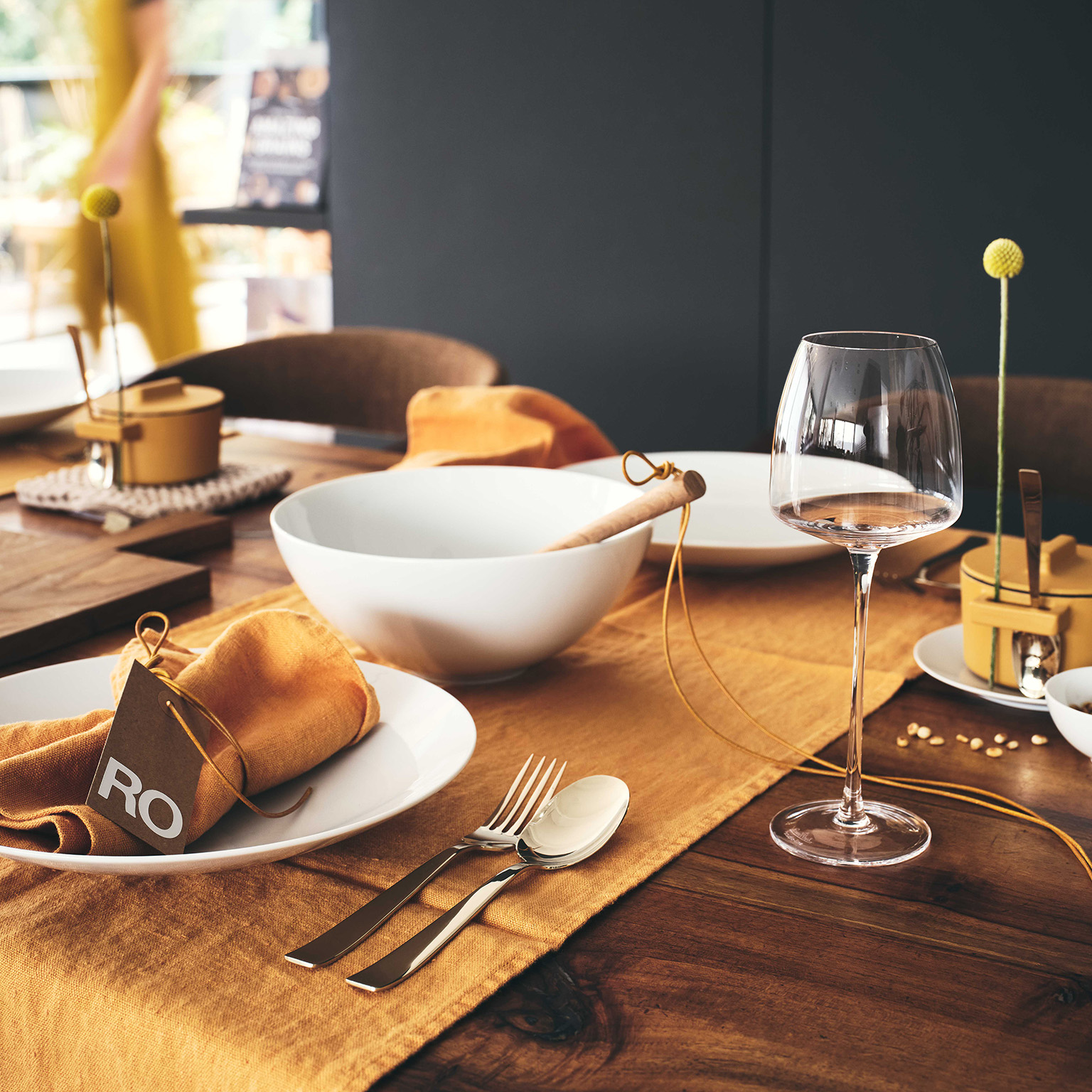 Close-up of a wooden table set in TAC White with a mustard yellow tablecloth, wine glasses and the TAC Allround bowl in the middle of the table.